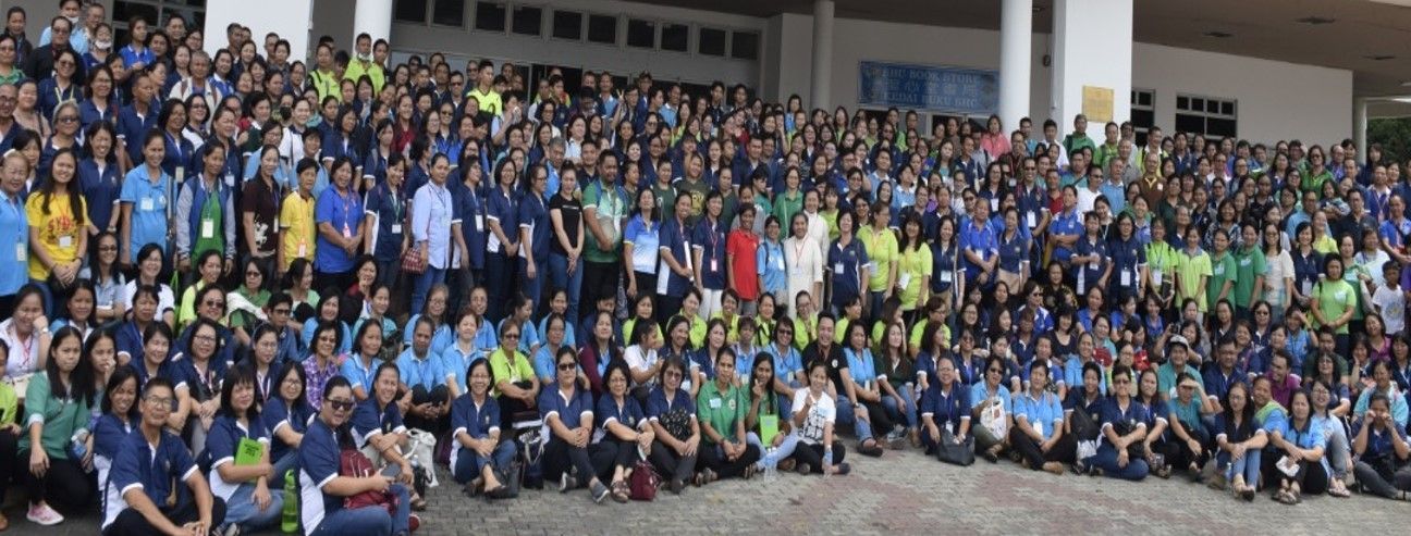 Large group of people in blue and green shirts, posing for a photo in front of a building.