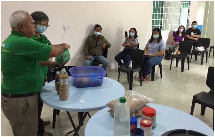 People in masks observing a presentation; two men in green shirts at a table with food items.