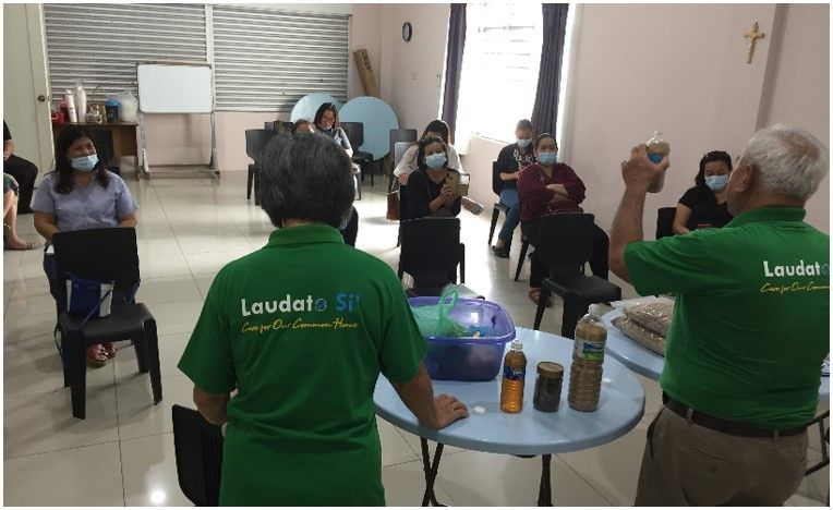 People in a room, wearing masks, listen to a presentation. Two people in green shirts stand by a table with bottles.