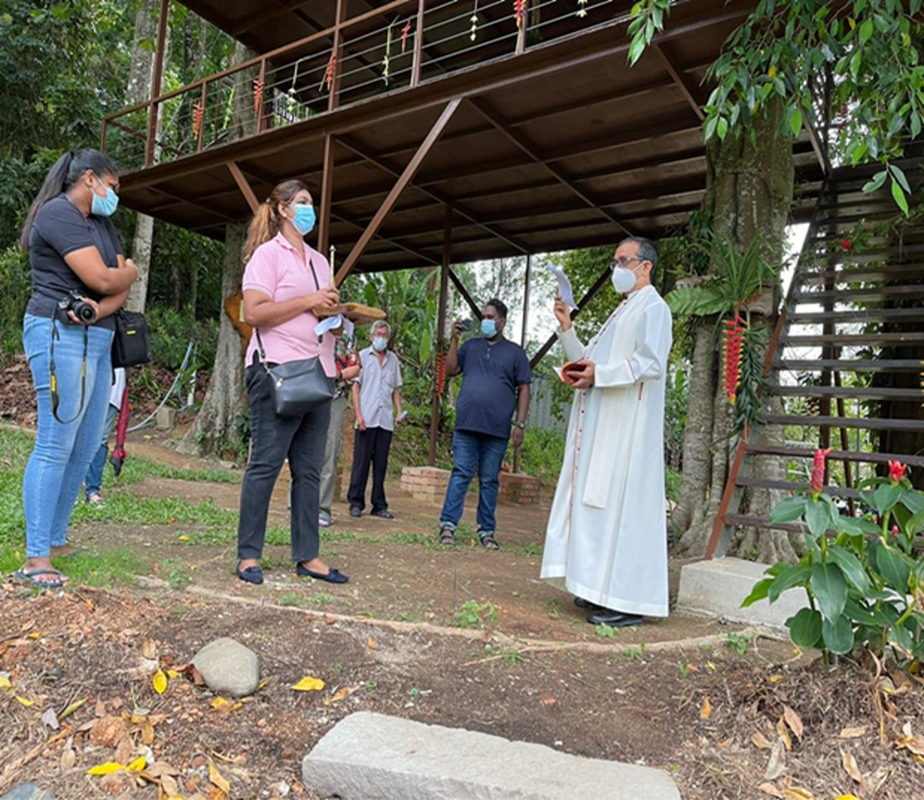 Group of people in masks stand outdoors as a robed person gestures, near a raised wooden platform.