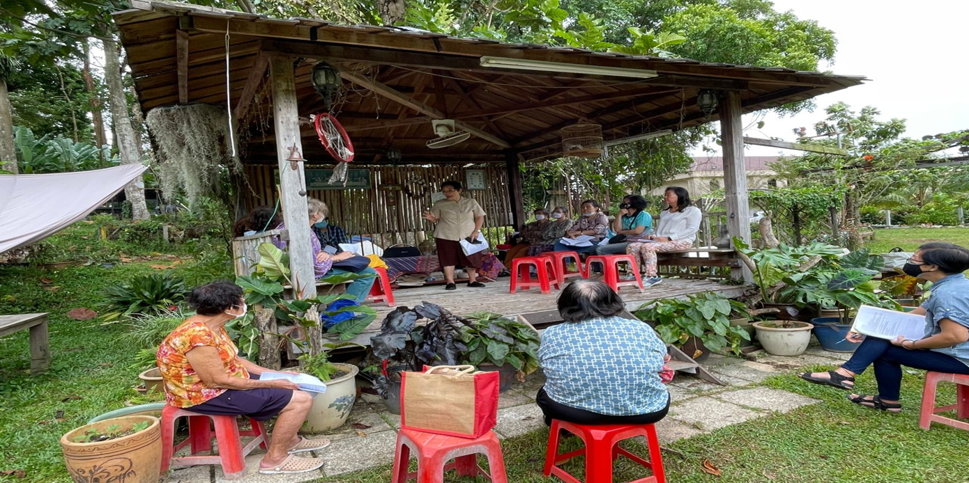 A group of people sitting under a shaded structure, some with papers, in an outdoor setting.
