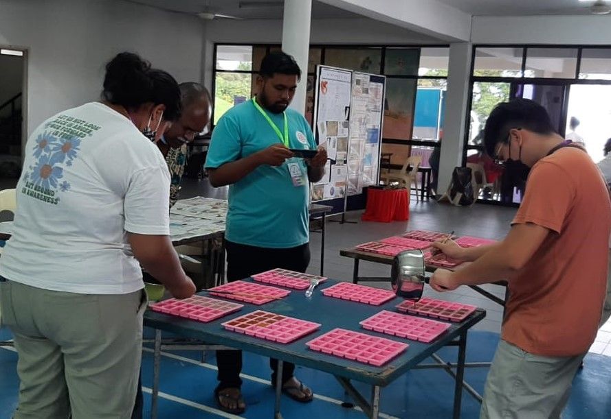 People working on a table with pink square objects. A man takes a picture. Indoors, open space.