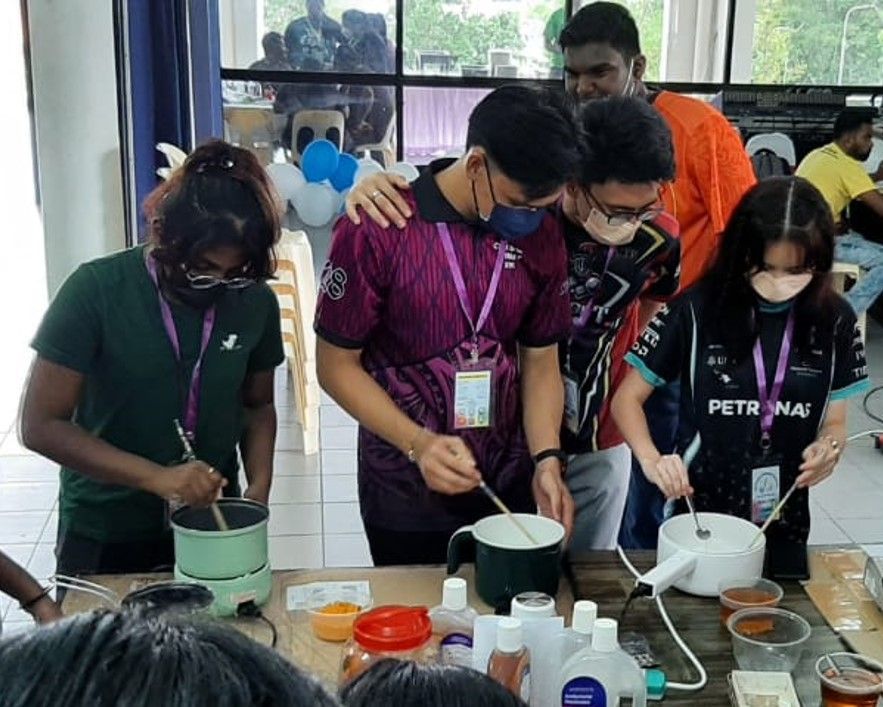 Four people stirring ingredients in pots at a table; one person has an arm on another's shoulder.
