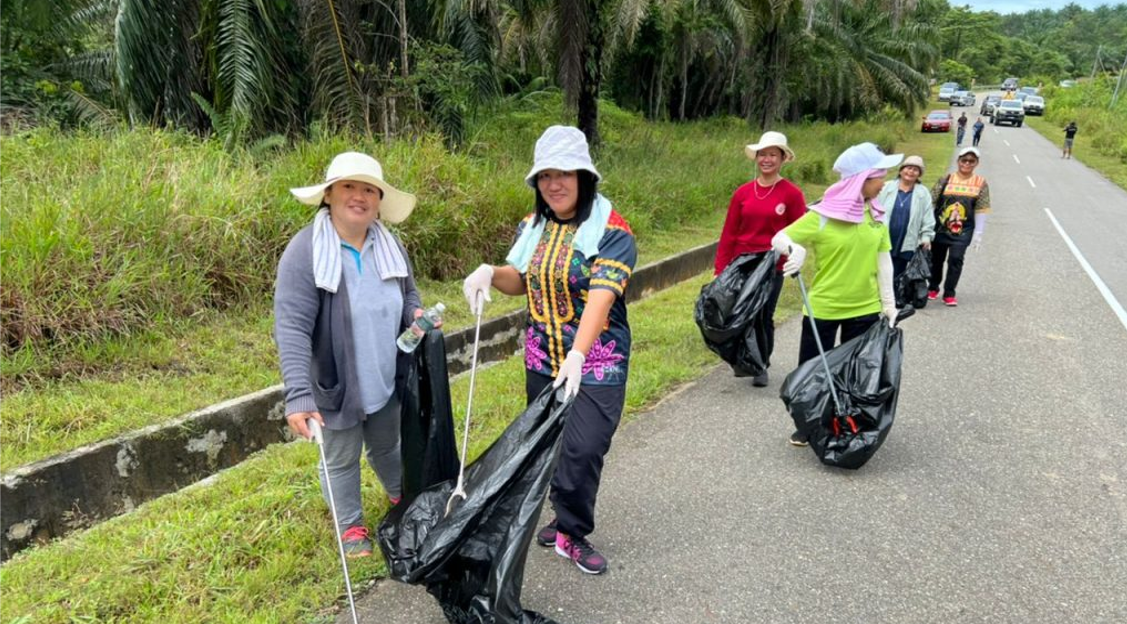 Volunteers collect trash along a roadside. They wear hats and carry black bags. Lush greenery and a gray road are in view.