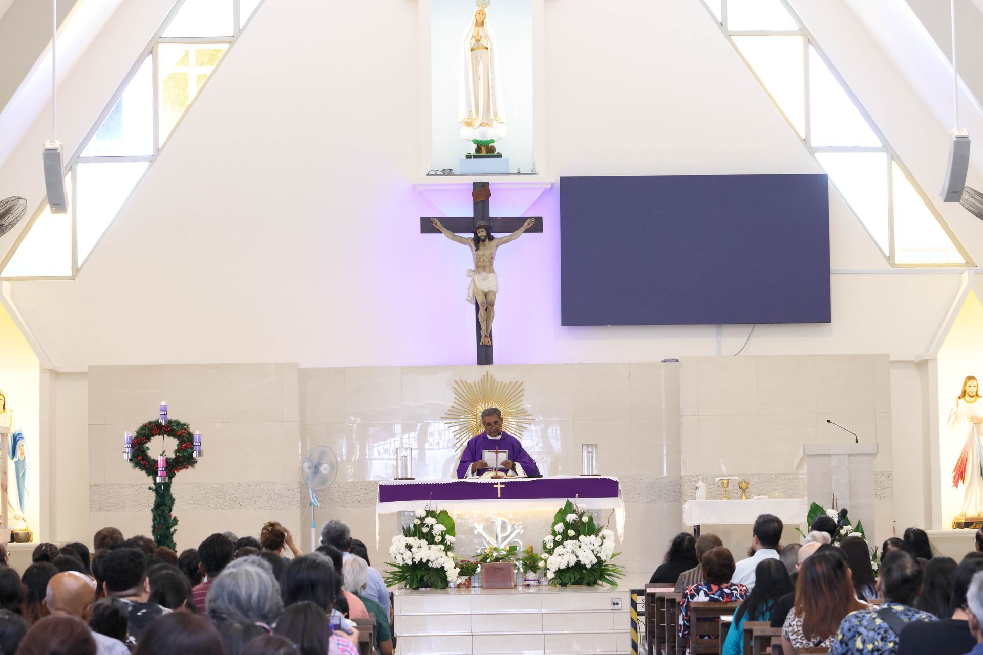 Priest at altar giving Mass in a church, facing a congregation. Crucifix and flowers are on the altar.