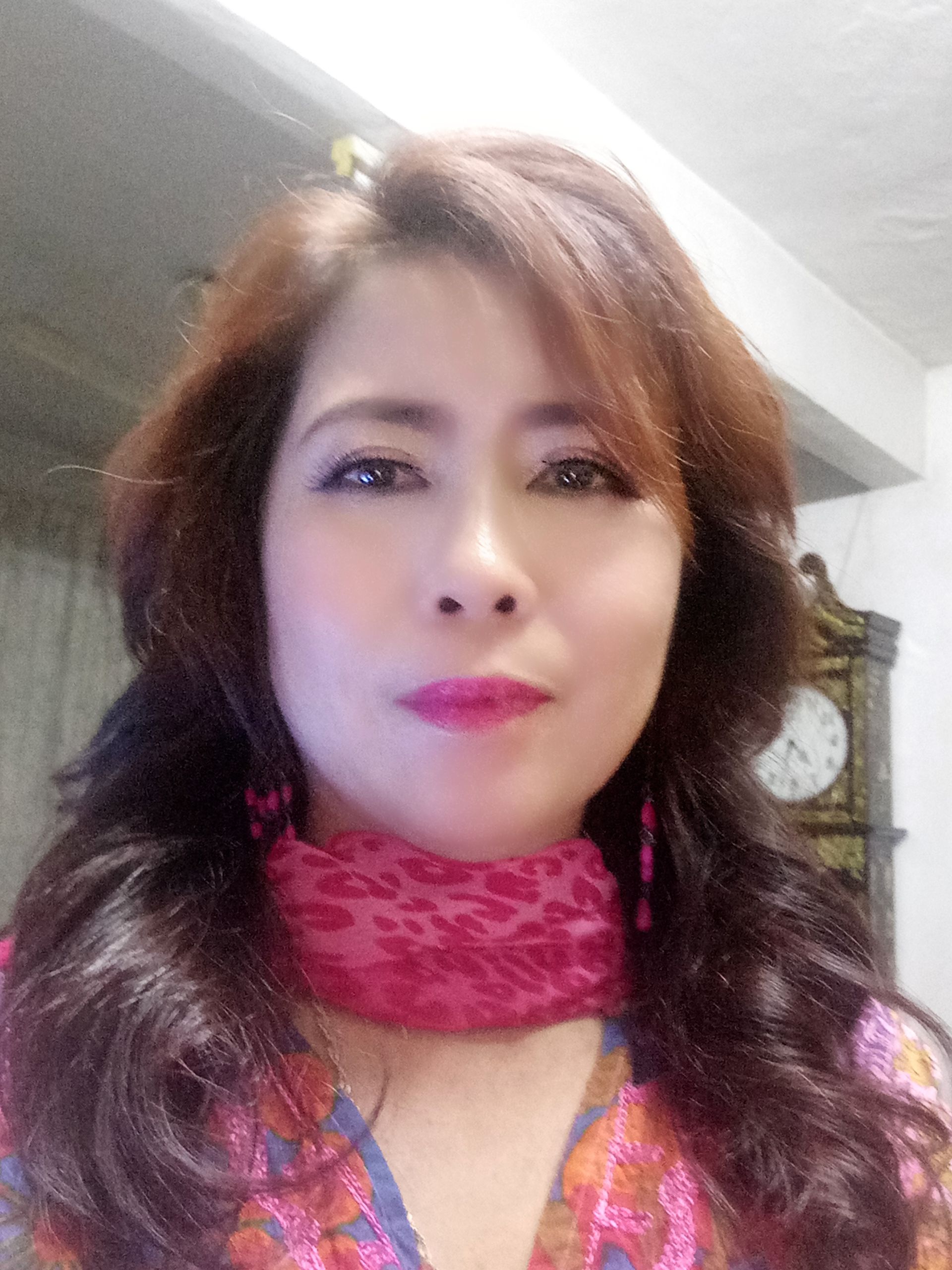 Woman with long, wavy brown hair, wearing a pink patterned scarf and top; clock in background.