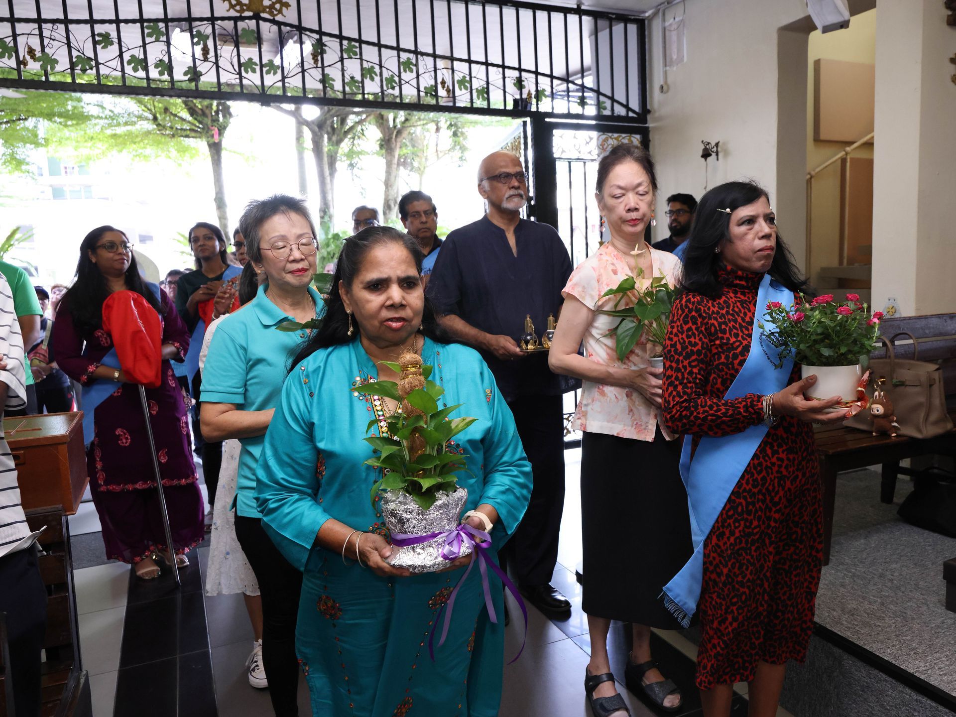 People carrying potted plants, walking indoors. Some are smiling, others are serious. Brightly lit entrance.