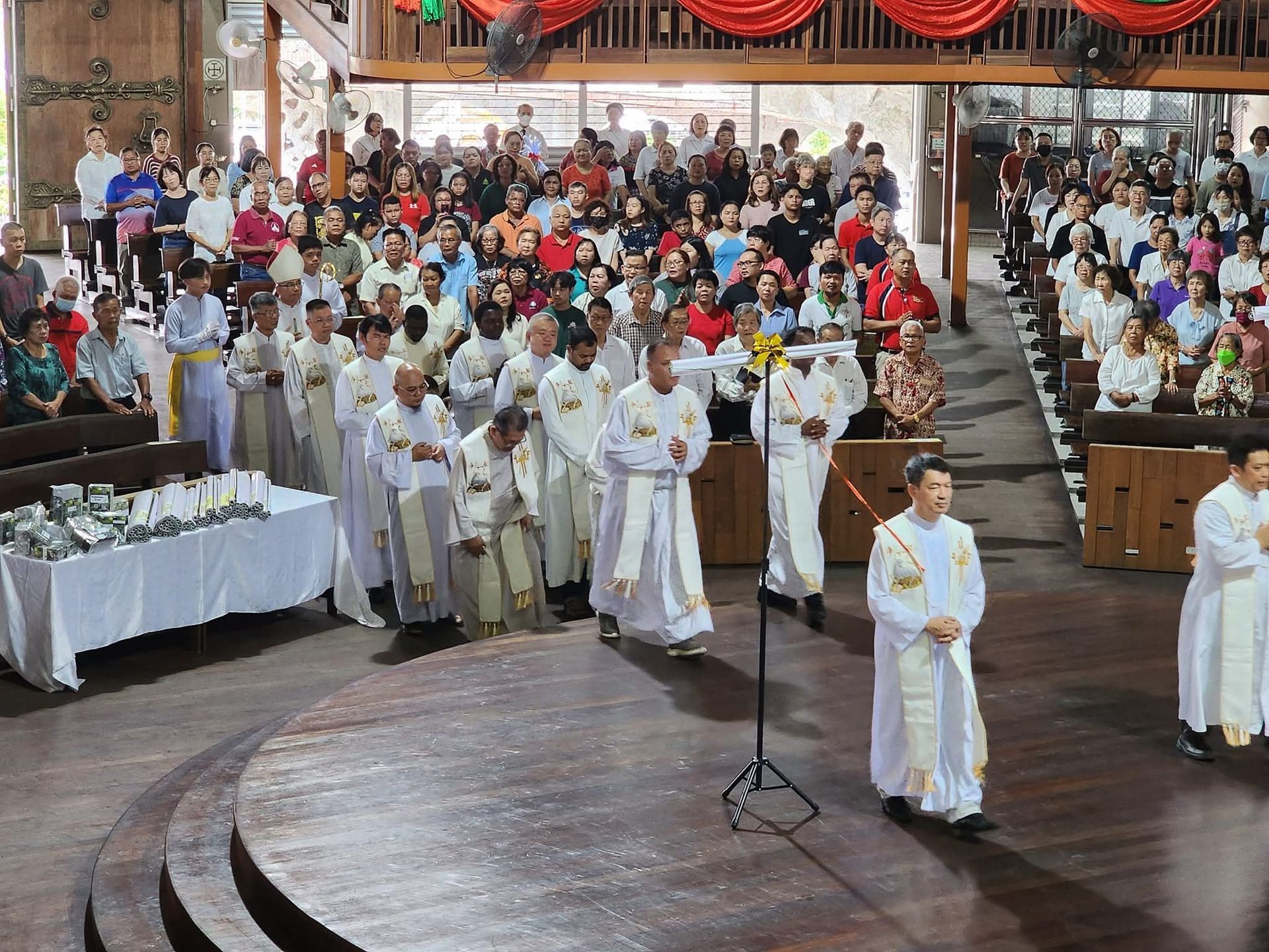 Clergy in white robes lead a procession through a church, with a large crowd in the background.
