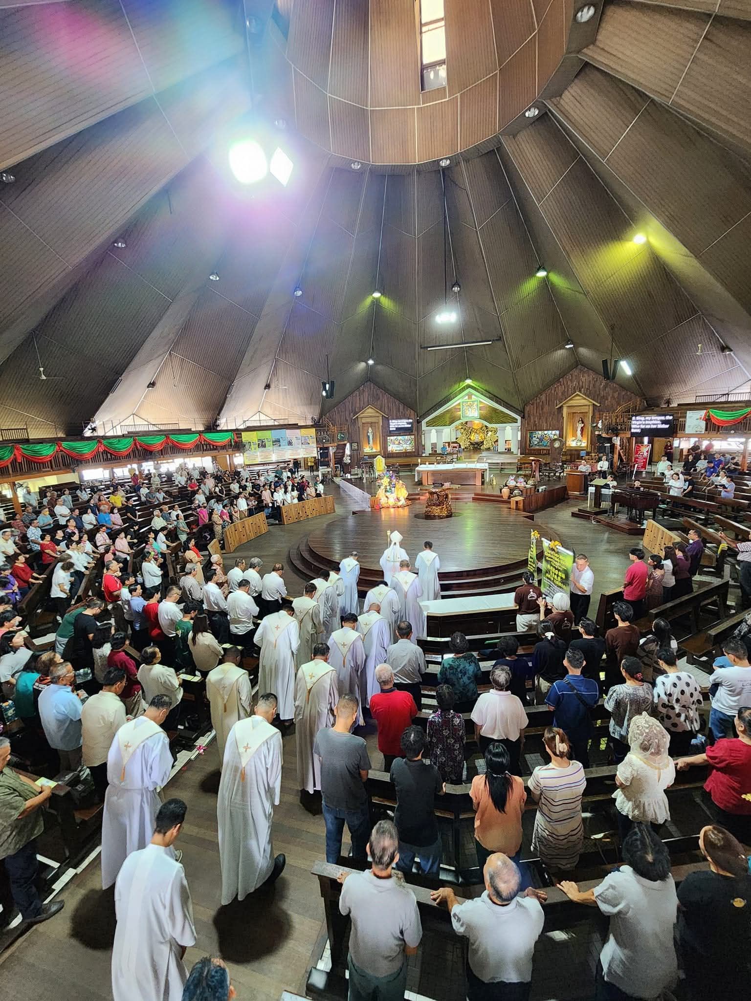 Interior view of a crowded church service. People seated and standing, a choir, and clergy at the altar.