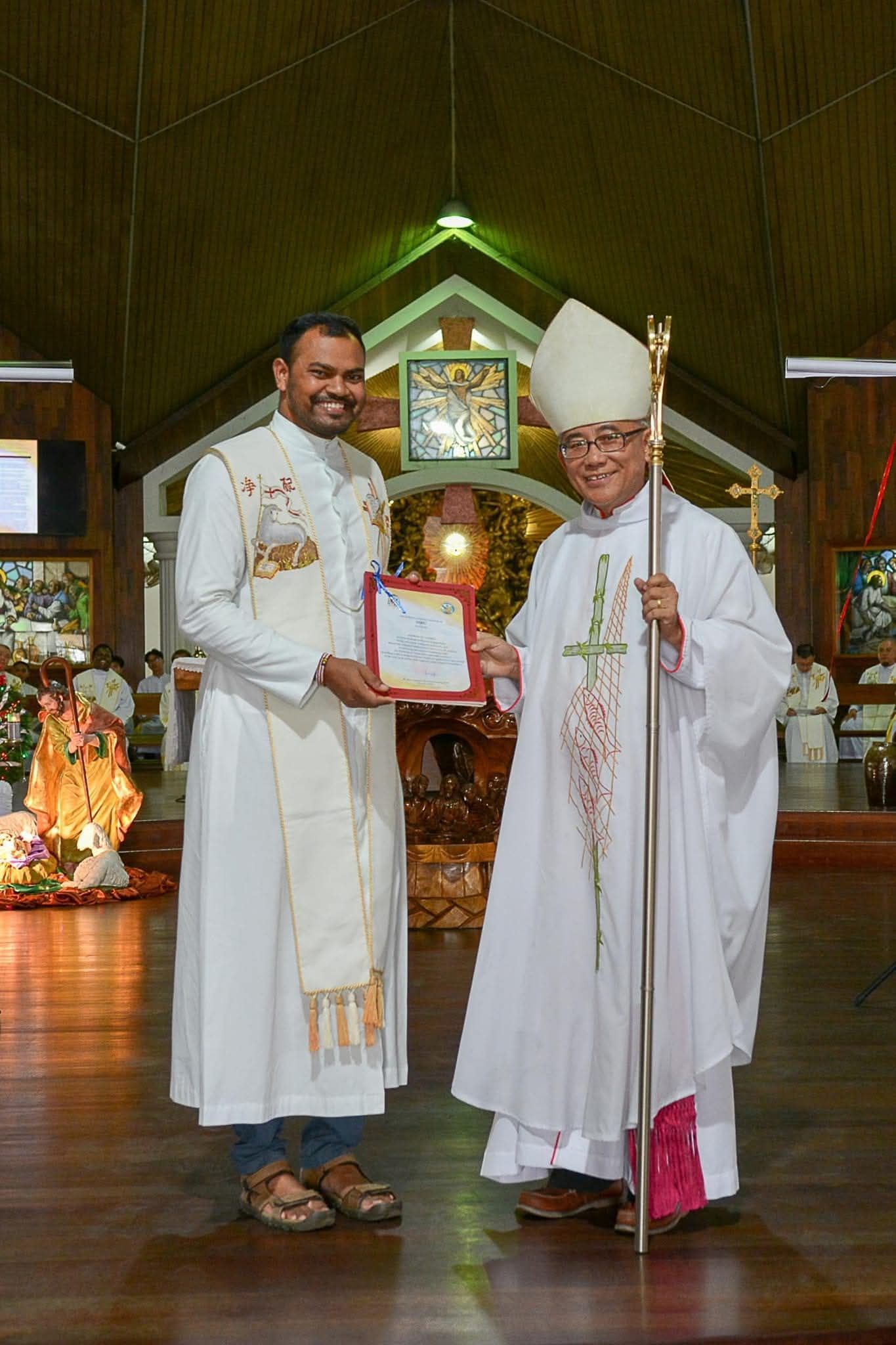 Cleric receives certificate from bishop in church. They are smiling. Indoor with lights.