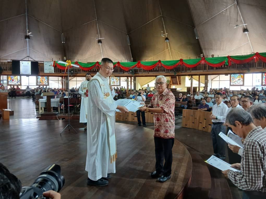 Priest handing document to a man in a church during a ceremony. Others hold papers and watch.