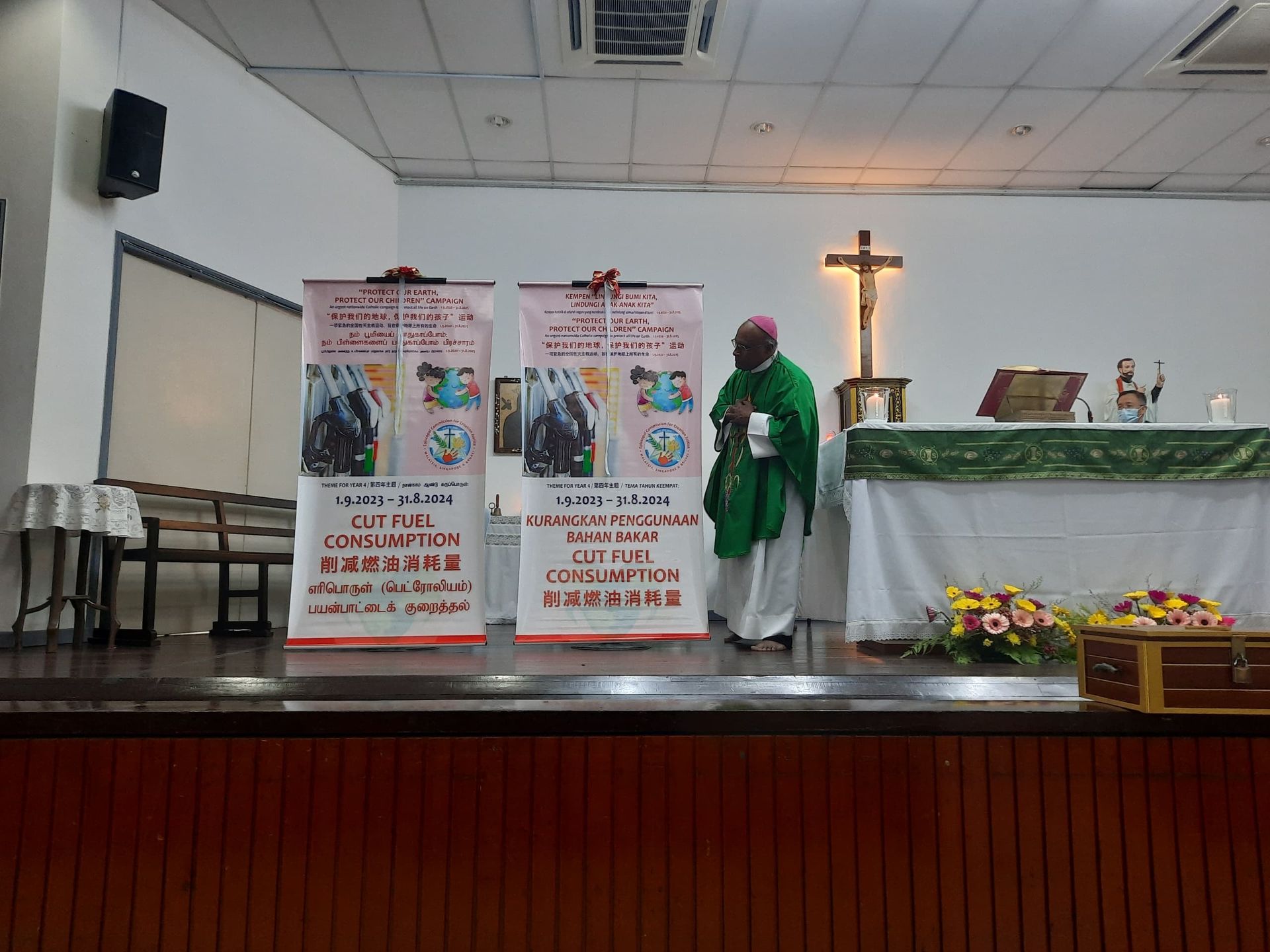 A bishop in green robes stands on a stage with banners and a cross, likely at a church event.