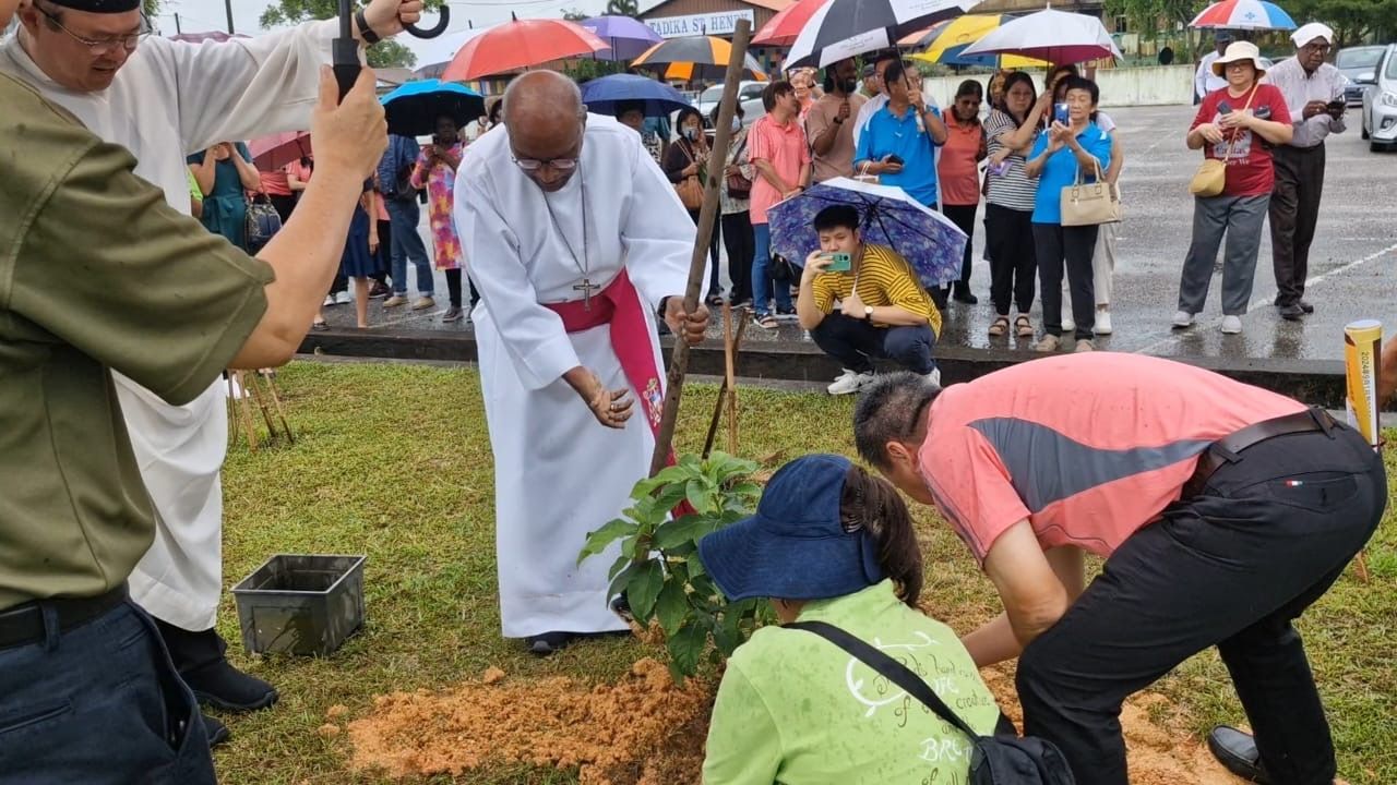 Clergy member plants a tree with assistance. Others watch, holding umbrellas in the rainy outdoor setting.