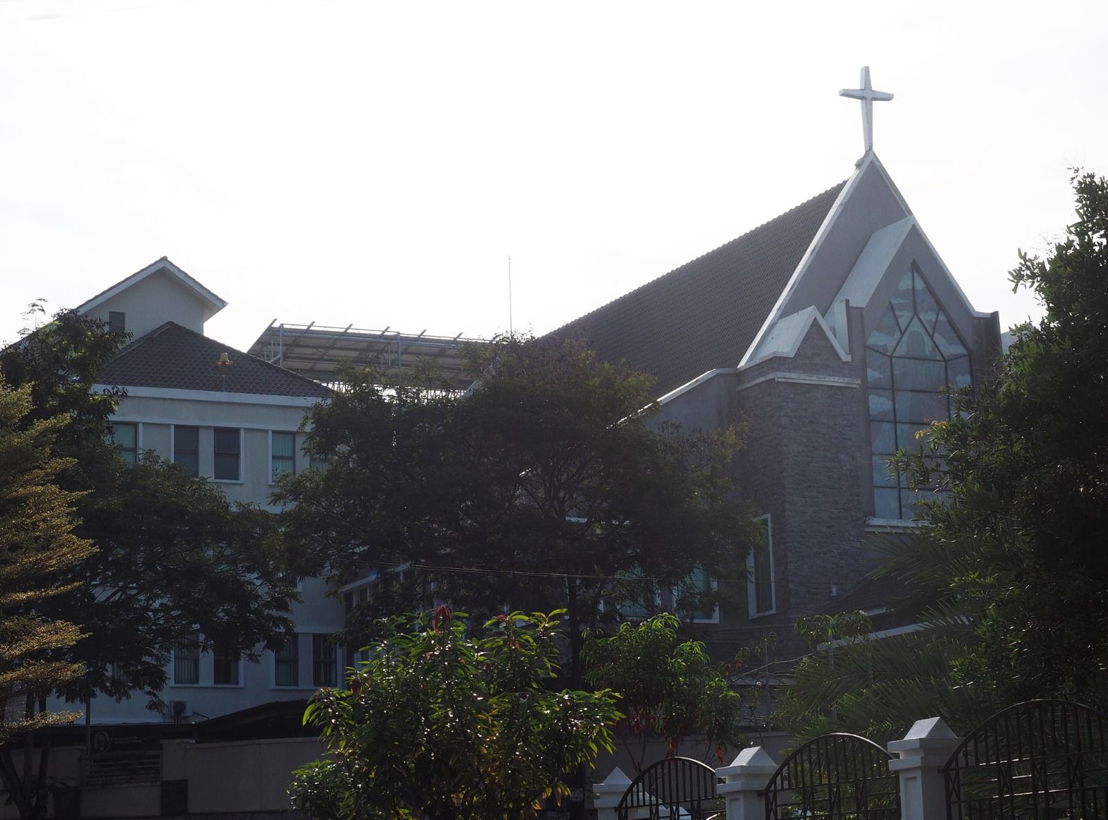 Church building with a cross on top, surrounded by trees and a white fence.