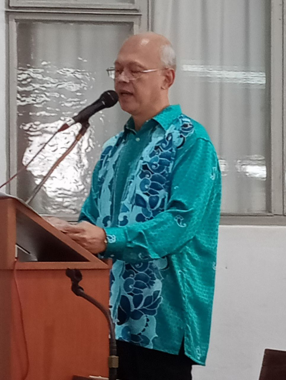 Man in turquoise patterned shirt speaking at a podium, indoors.
