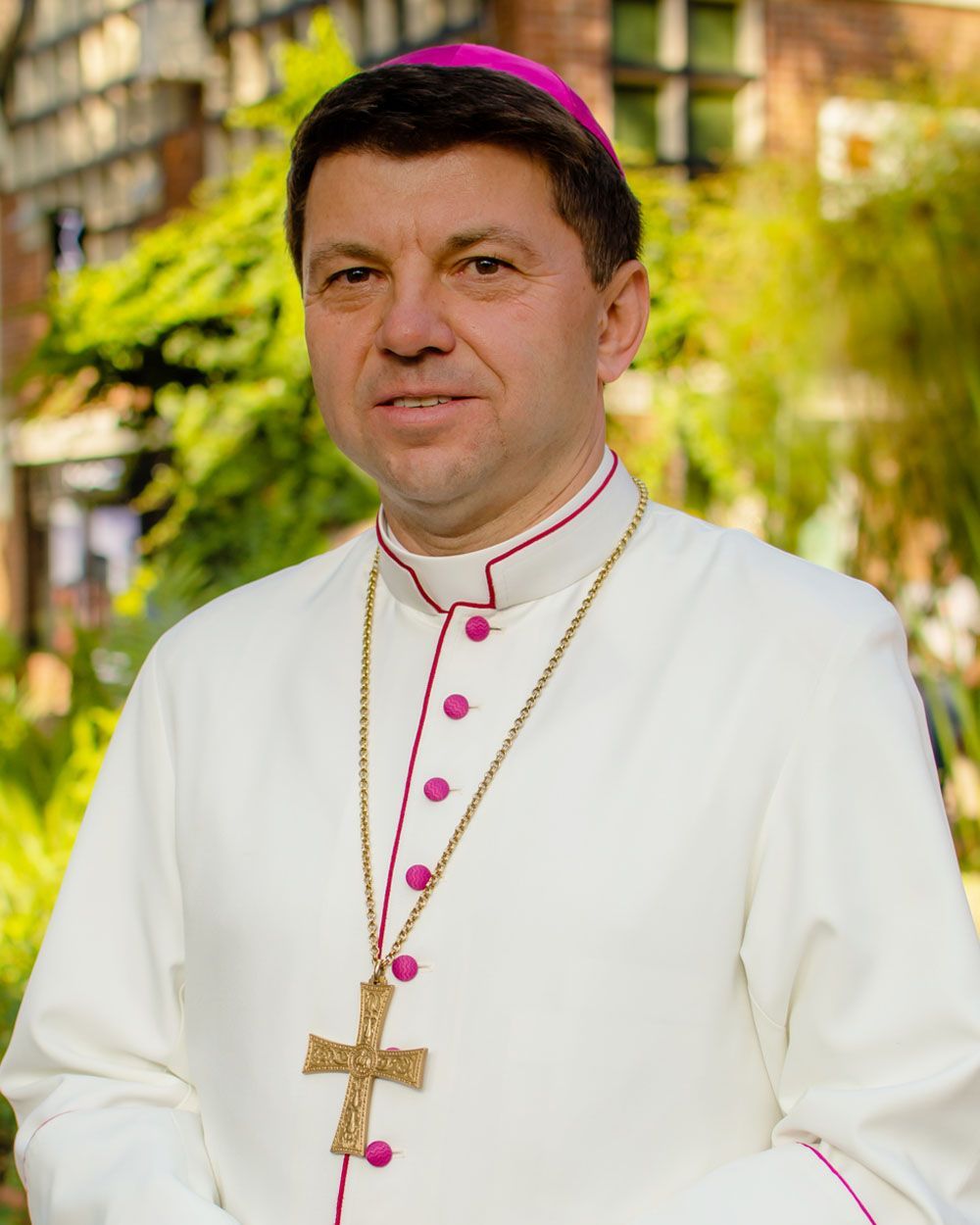 Man in white clerical robes, pink trim, and cross necklace. Outdoors, background of greenery and building.