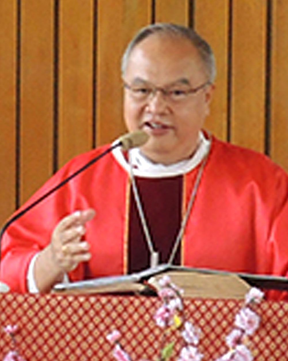 Clergyman speaking at a podium, wearing a red robe, white collar, and glasses.