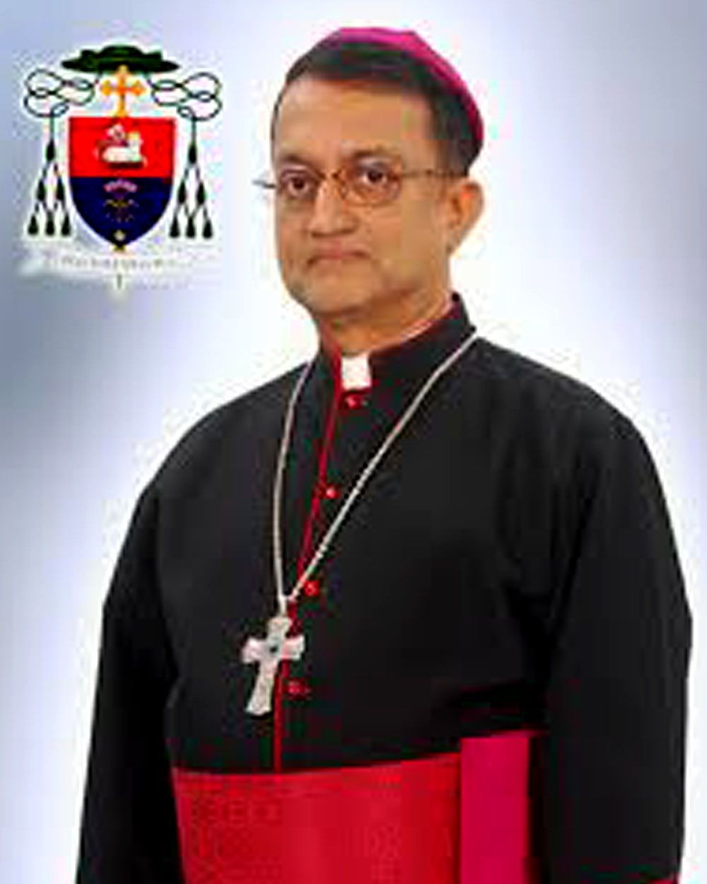 Bishop in black cassock with red sash and zucchetto, wearing a cross necklace, and glasses. Official coat of arms above.