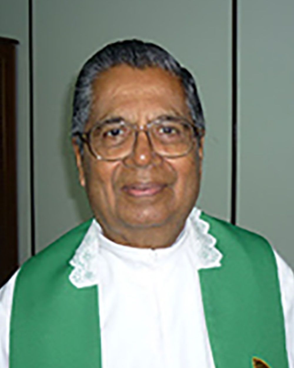 Clergyman wearing glasses, a green stole, and a white collar, smiling indoors.