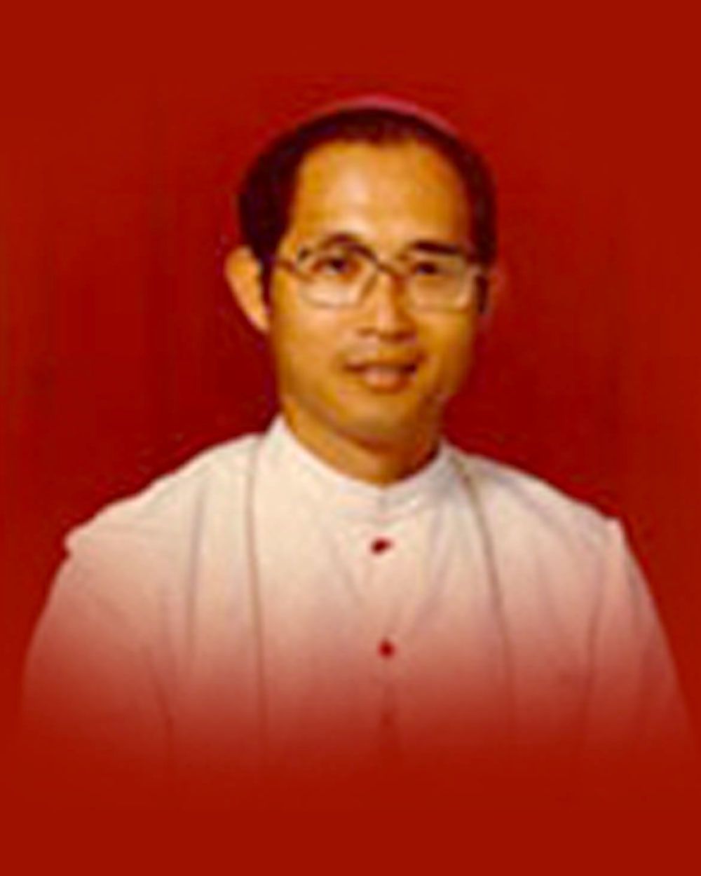 Smiling man in white clerical collar, glasses, against red backdrop.