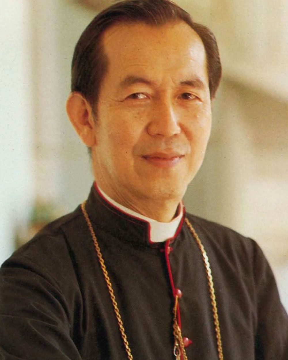Clergyman in black clerical attire, red trim, gold chain, smiling, head and shoulders portrait.