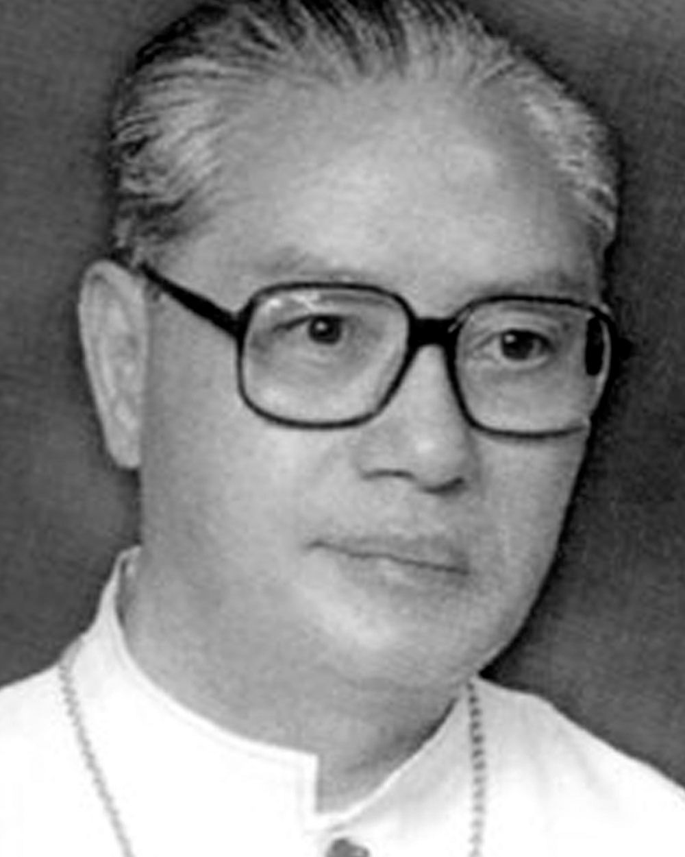 Portrait of an older man wearing glasses, clerical collar, and a serious expression.
