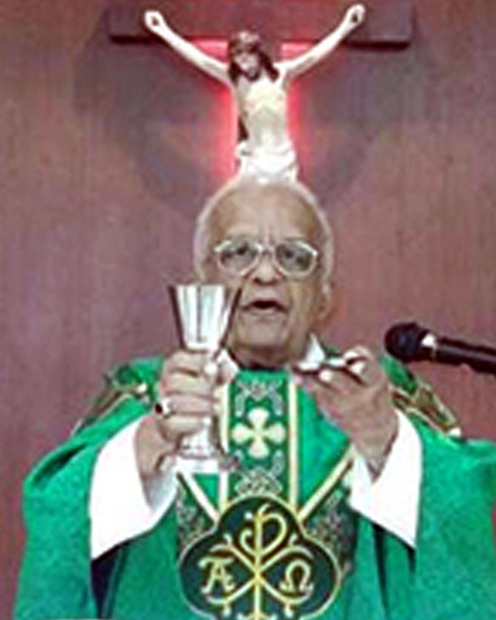 A priest holding a chalice and a wafer during a religious ceremony, with a crucifix in the background.