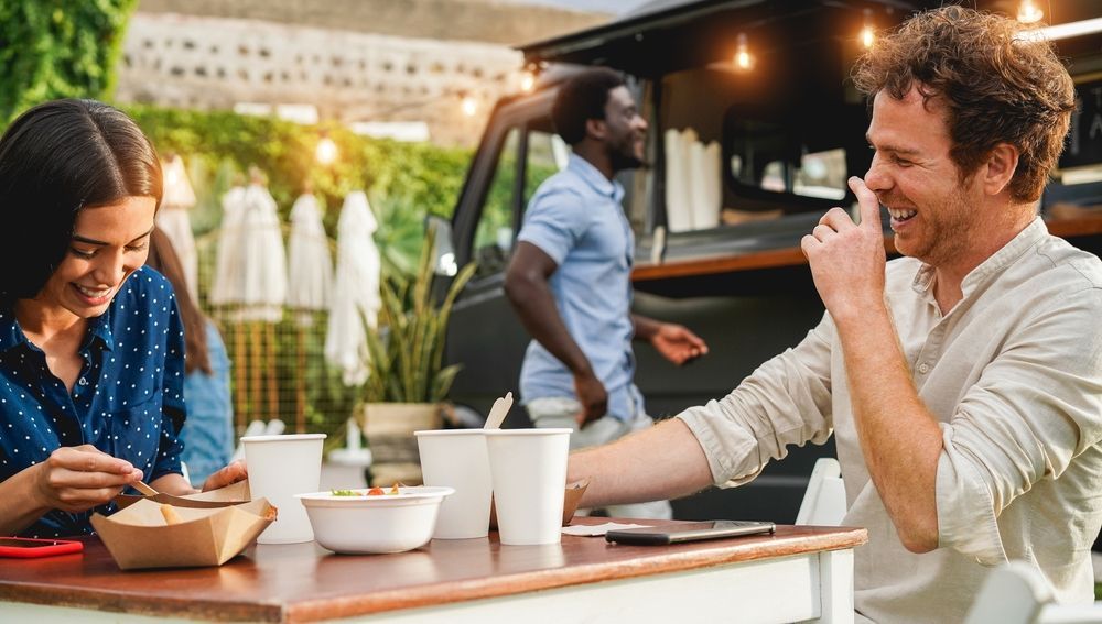 Two people eating and laughing at a table in front of a food truck. A server walks by. The setting is outdoors.