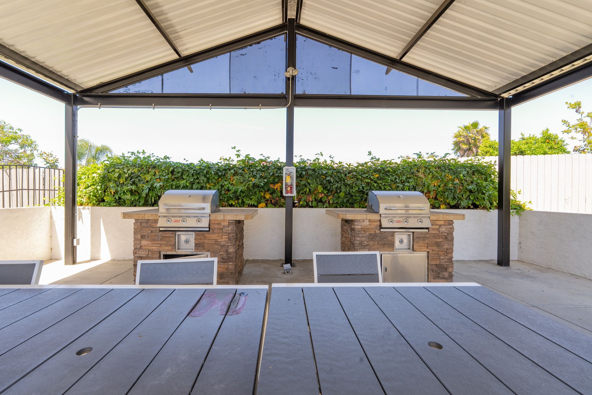 Covered outdoor patio with wooden tables, built-in stone grills, and benches against a hedge backdrop
