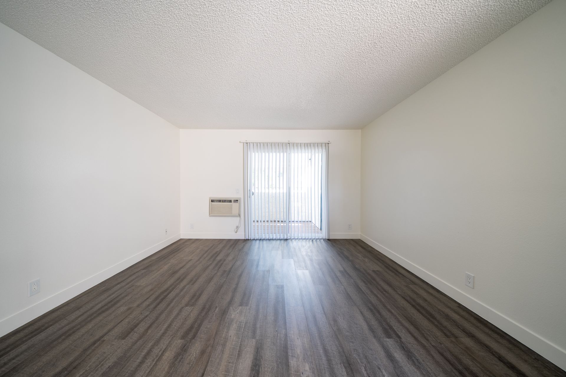 Empty room with gray wood floors, white walls, and sliding glass doors with vertical blinds