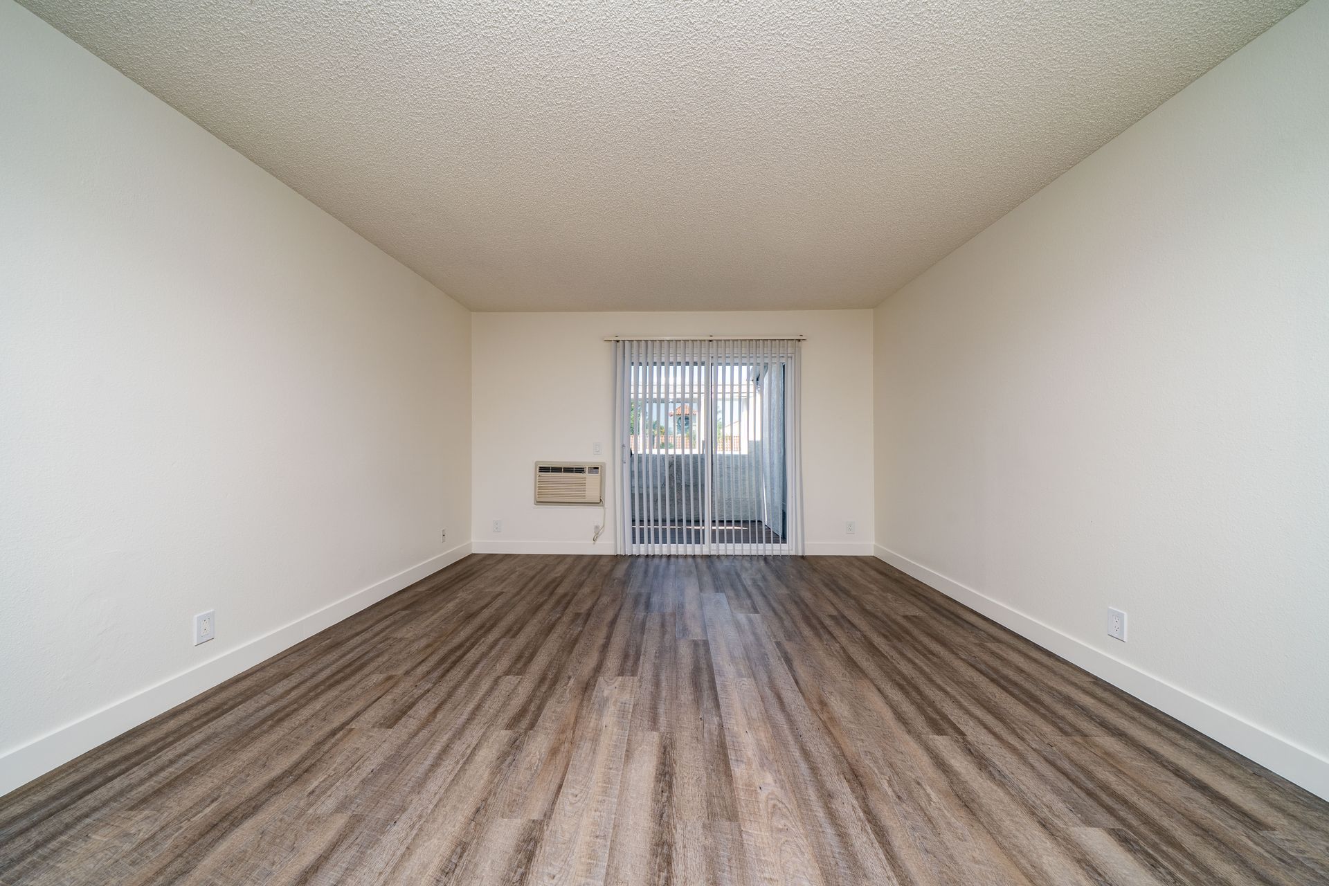Empty living room with wood-look flooring, white walls, and sliding glass door with vertical blinds.