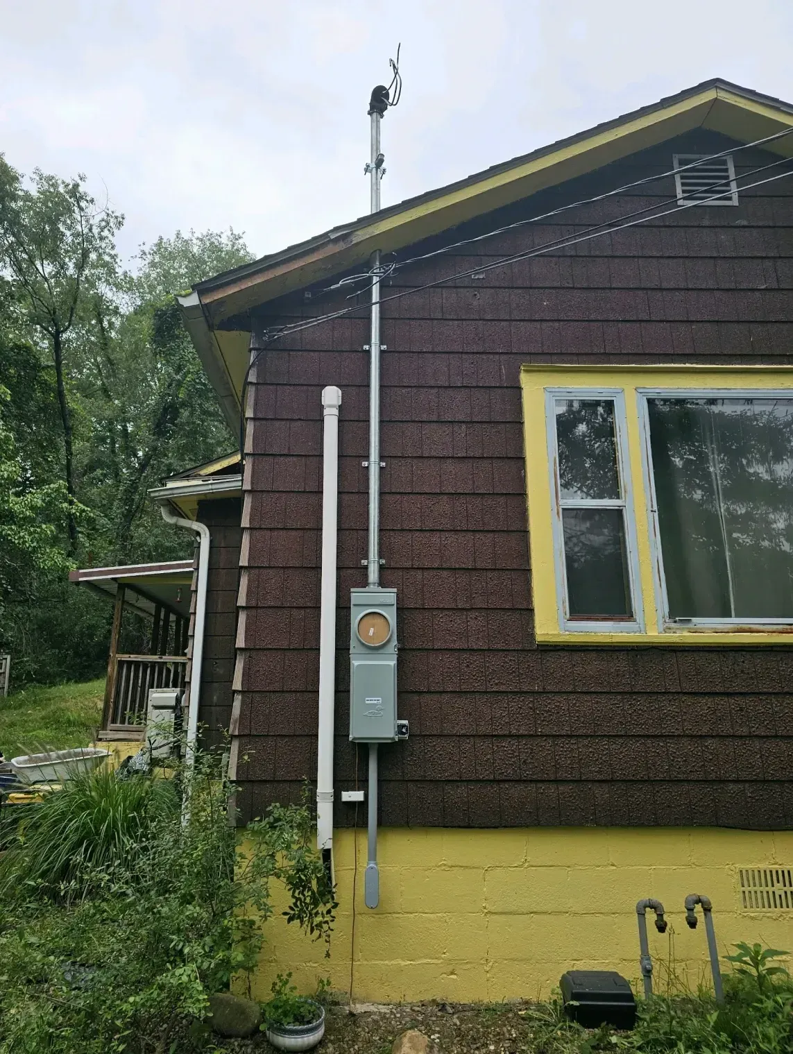 A residential exterior wall with dark brown shingles and a yellow foundation, featuring a gray electrical meter box and mast.