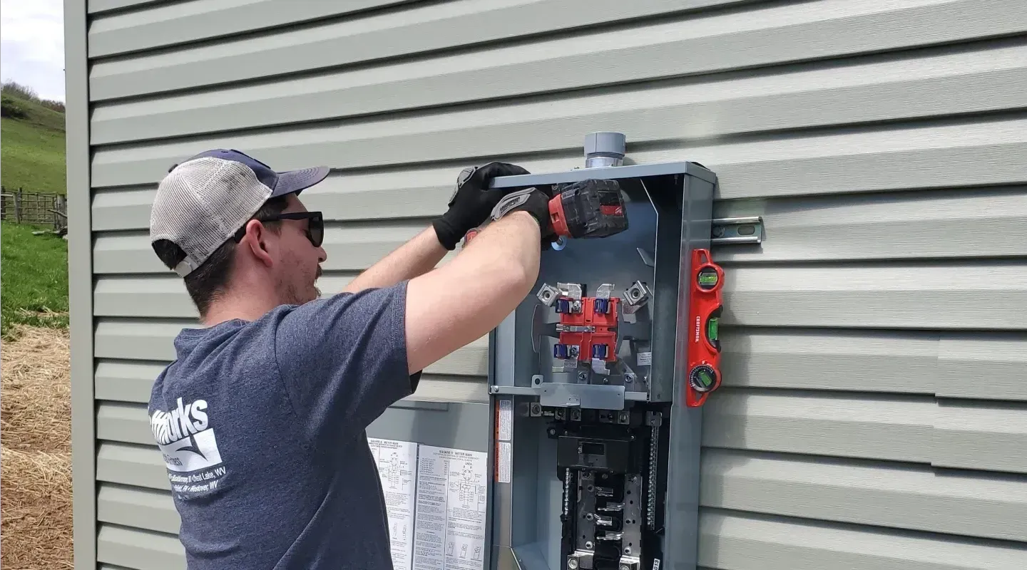 A person in a baseball cap and work shirt installs an electrical meter box onto an outdoor vinyl-sided wall.