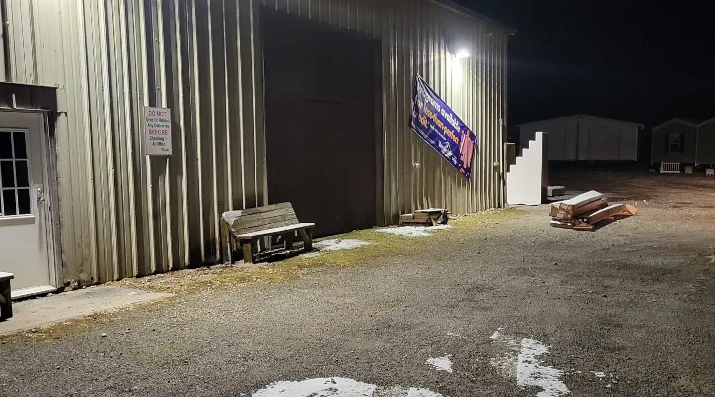 Exterior night view of a corrugated metal warehouse with a lit doorway, signage, a bench, and logs on a gravel lot.