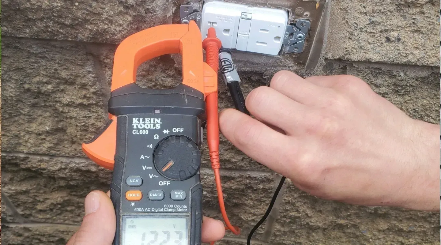 A person uses a digital multimeter to test voltage at an outdoor wall electrical outlet.