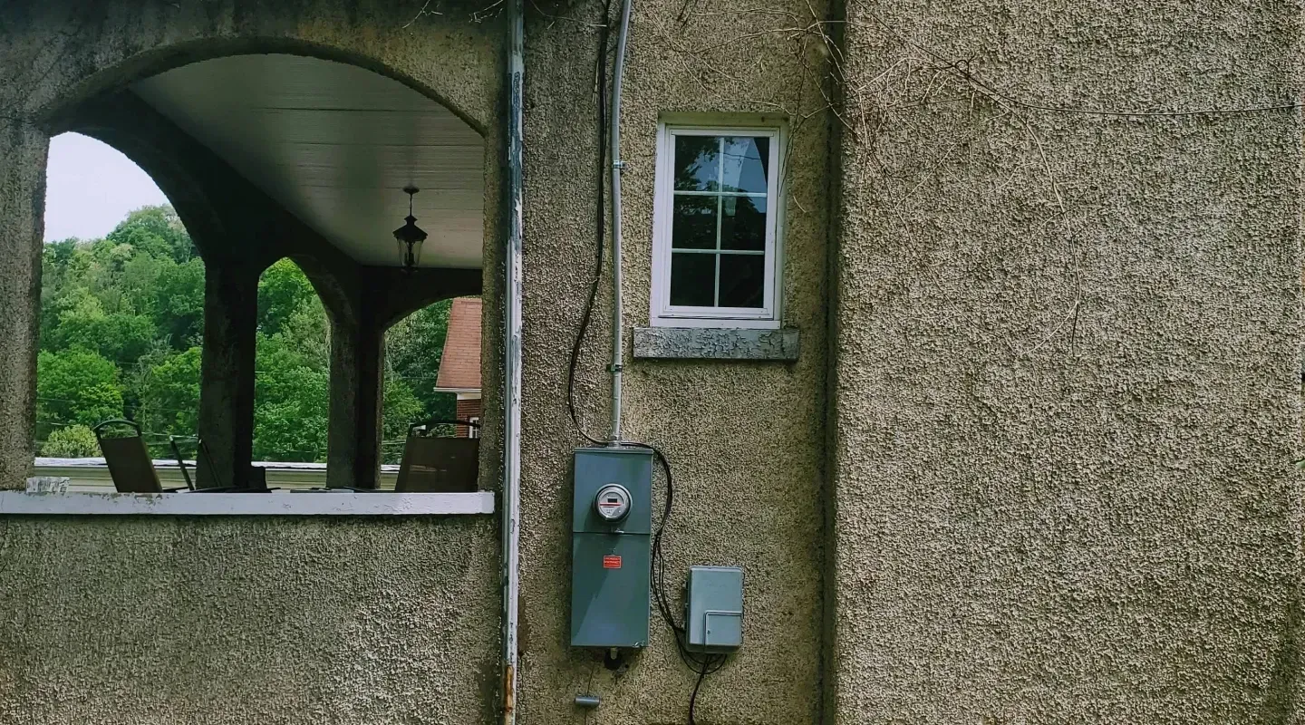 A pebble-dash building exterior with an arched porch on the left, a small window, and electrical equipment on the wall.
