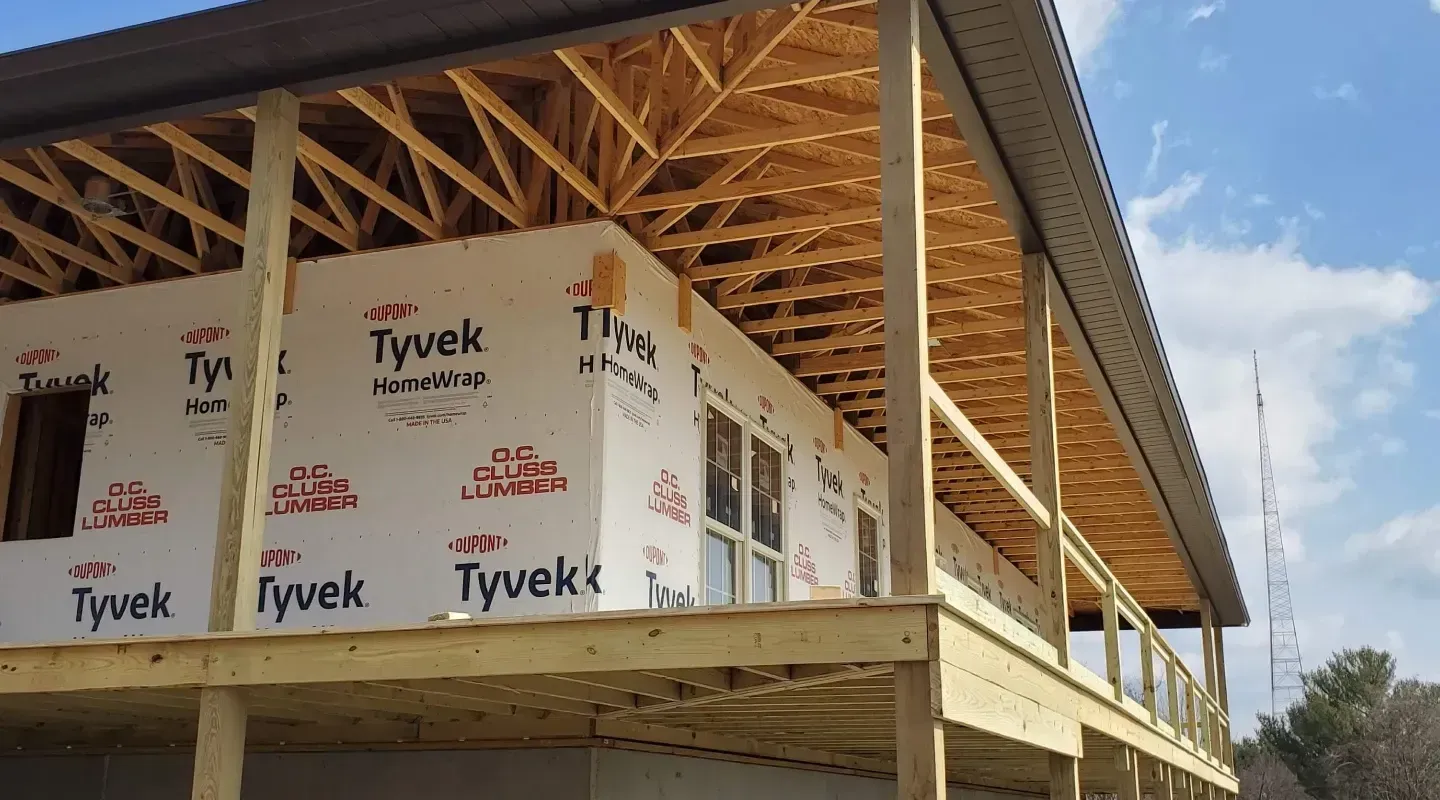 Exterior view of a residential building under construction with exposed wooden roof trusses, deck frame, and Tyvek wrap.
