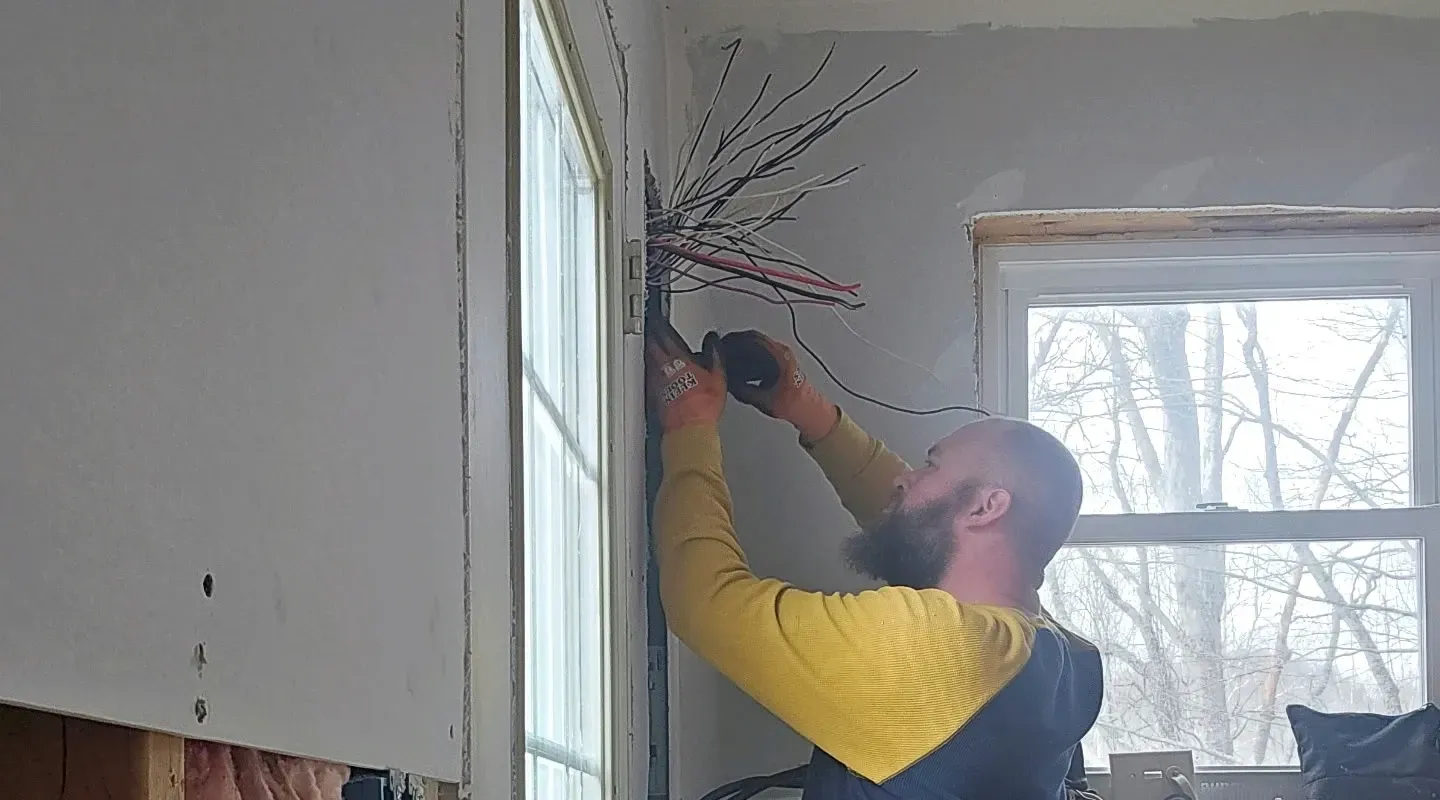 A person in a yellow and dark blue long-sleeved shirt installs electrical wiring in a wall next to a window.