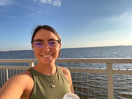 Smiling person taking a selfie by the water on a sunny day, wearing glasses and a green tank top