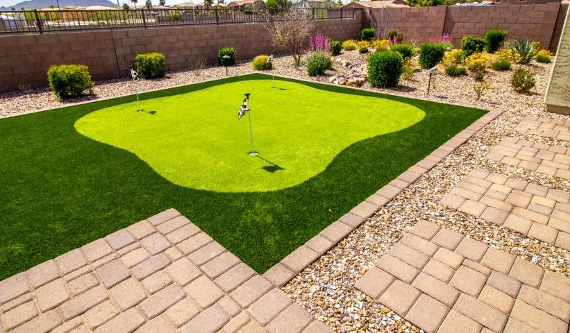 A putting green with two holes and flags, surrounded by artificial grass, stone pavers, and desert landscaping.