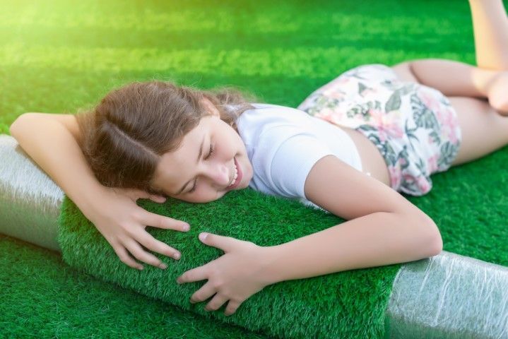 A person rests their head on a rolled piece of artificial turf while lying on a larger expanse of the same green material.