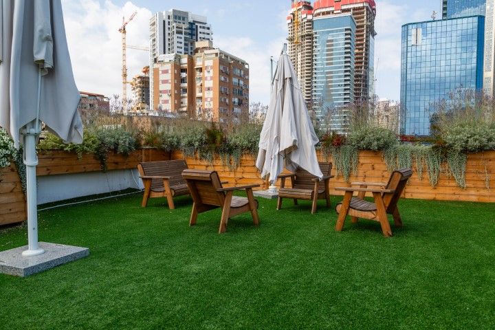 A rooftop terrace with artificial turf, wooden furniture, and umbrellas set against a city skyline with modern buildings.