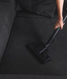 Hand holding a black vacuum nozzle over a dark carpeted sofa.