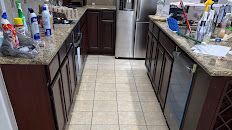 Narrow kitchen with granite counters, dark cabinets, tiled floor, and cleaning supplies on both sides.