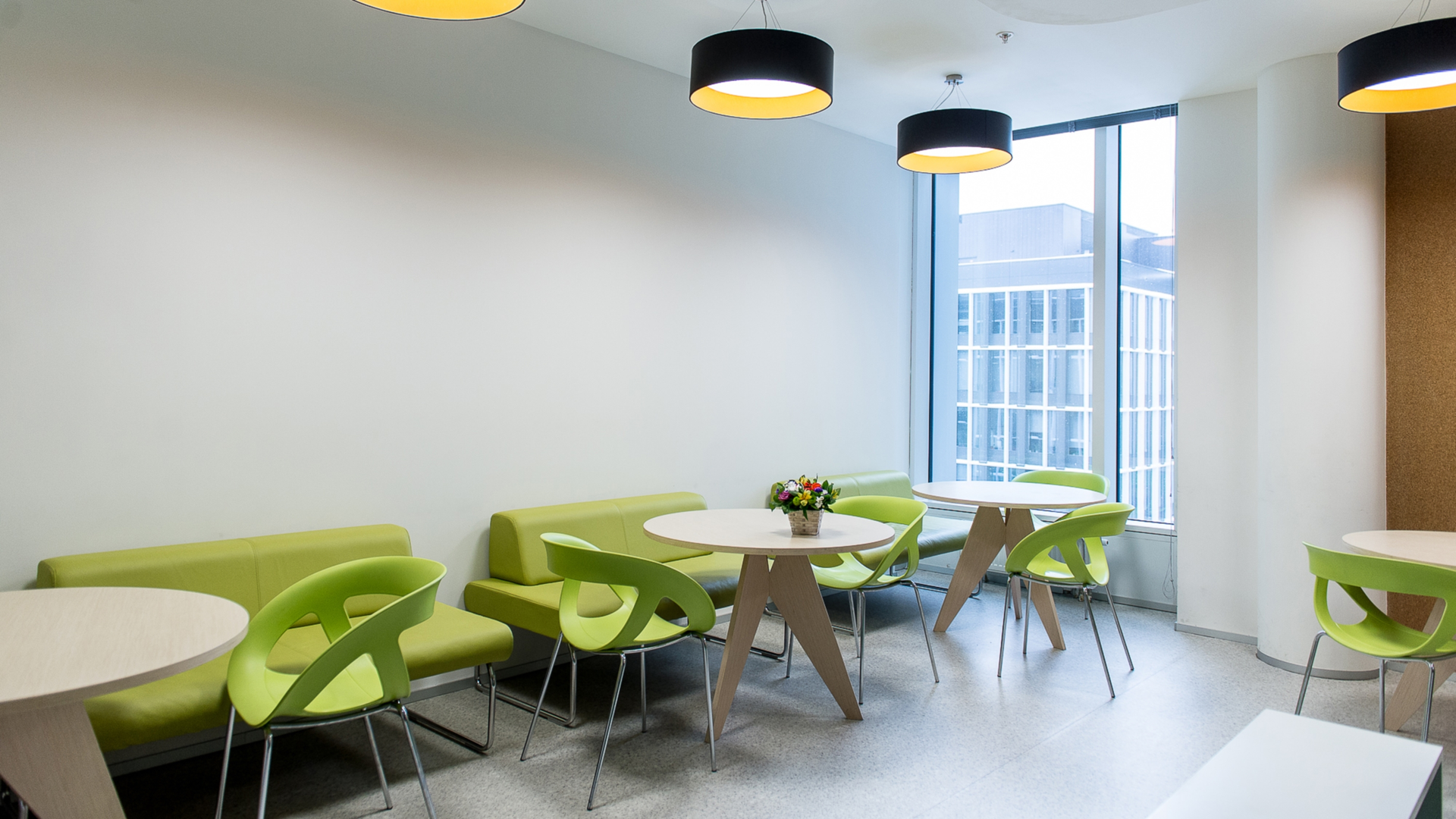 A modern office break room with light green booths, tables, lime green chairs, and black hanging lights by a window.