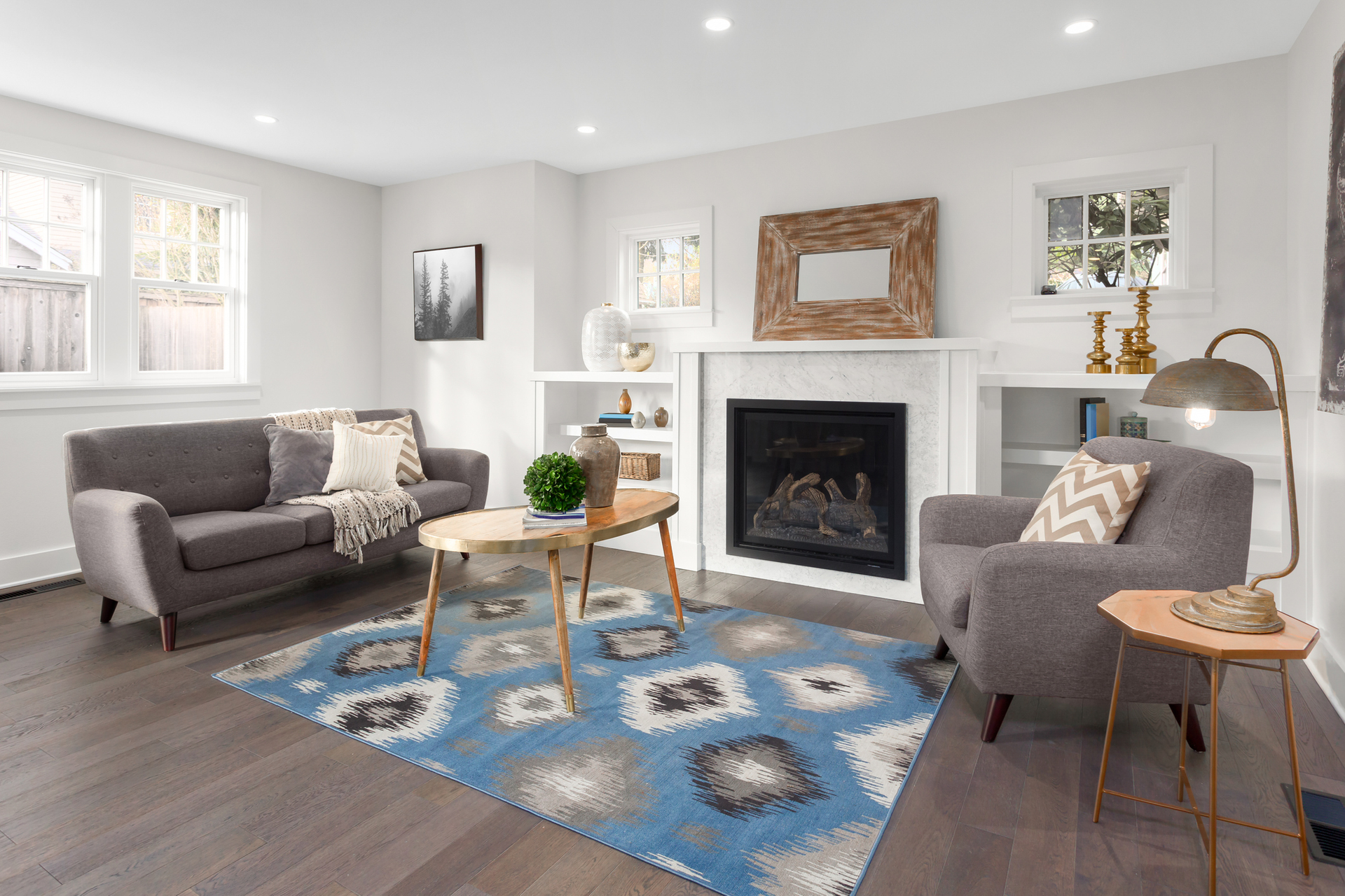 A modern living room with a grey sofa, matching armchair, blue patterned rug, and a fireplace with a wood-framed mirror.