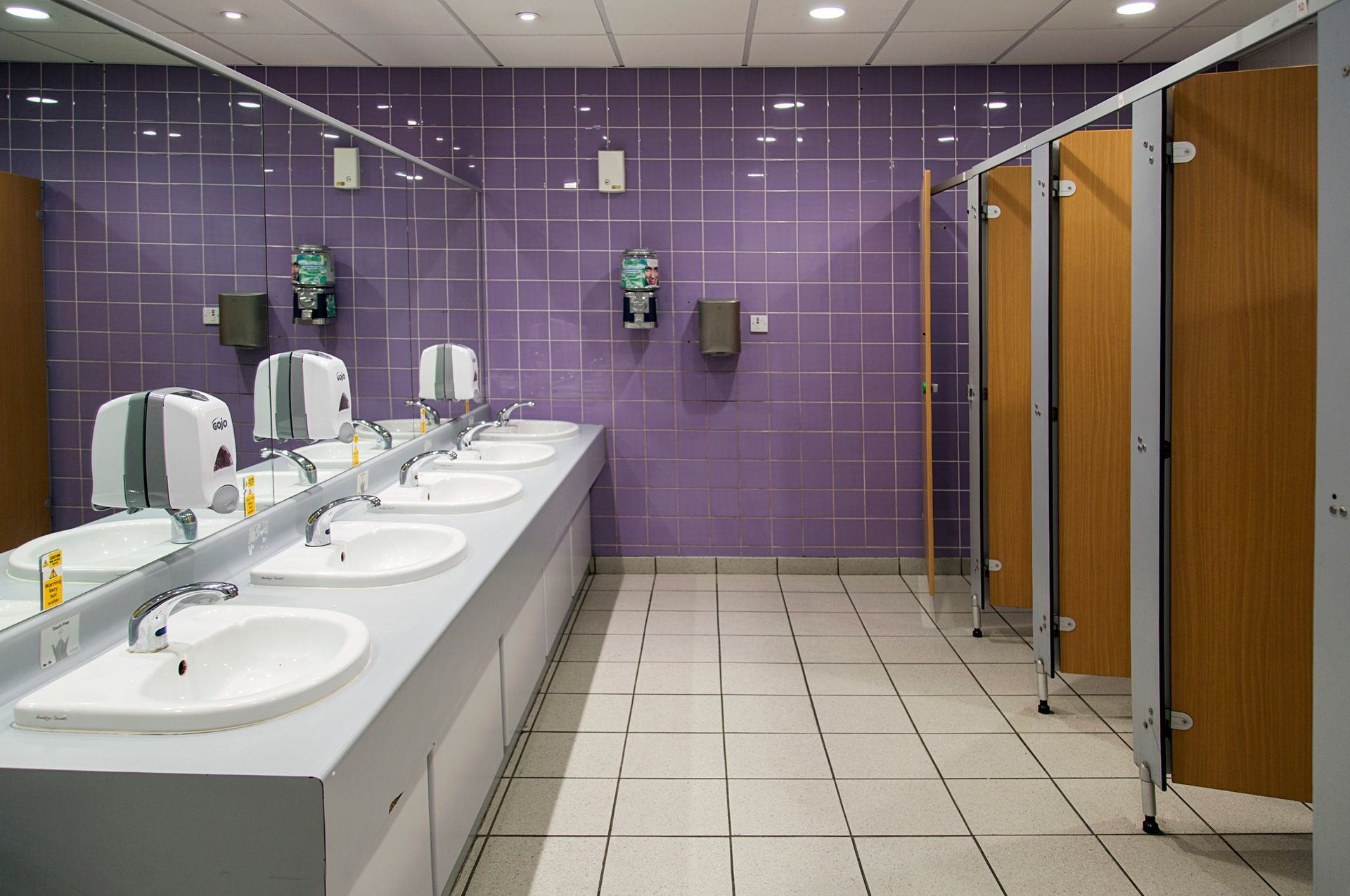 A modern public restroom with white sinks, chrome faucets, soap dispensers, purple tiled walls, and wood-paneled stalls.