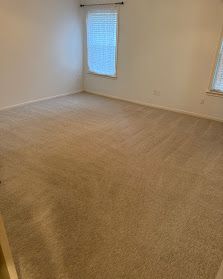 Empty beige-carpeted room with white walls and two windows letting in daylight.
