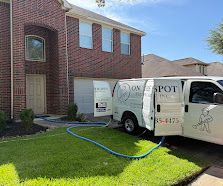 White van with hoses parked on a suburban lawn beside a brick house under clear blue sky.