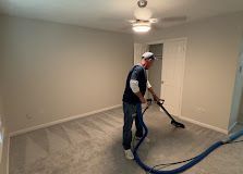Worker vacuuming carpet in an empty room with beige walls and ceiling fan